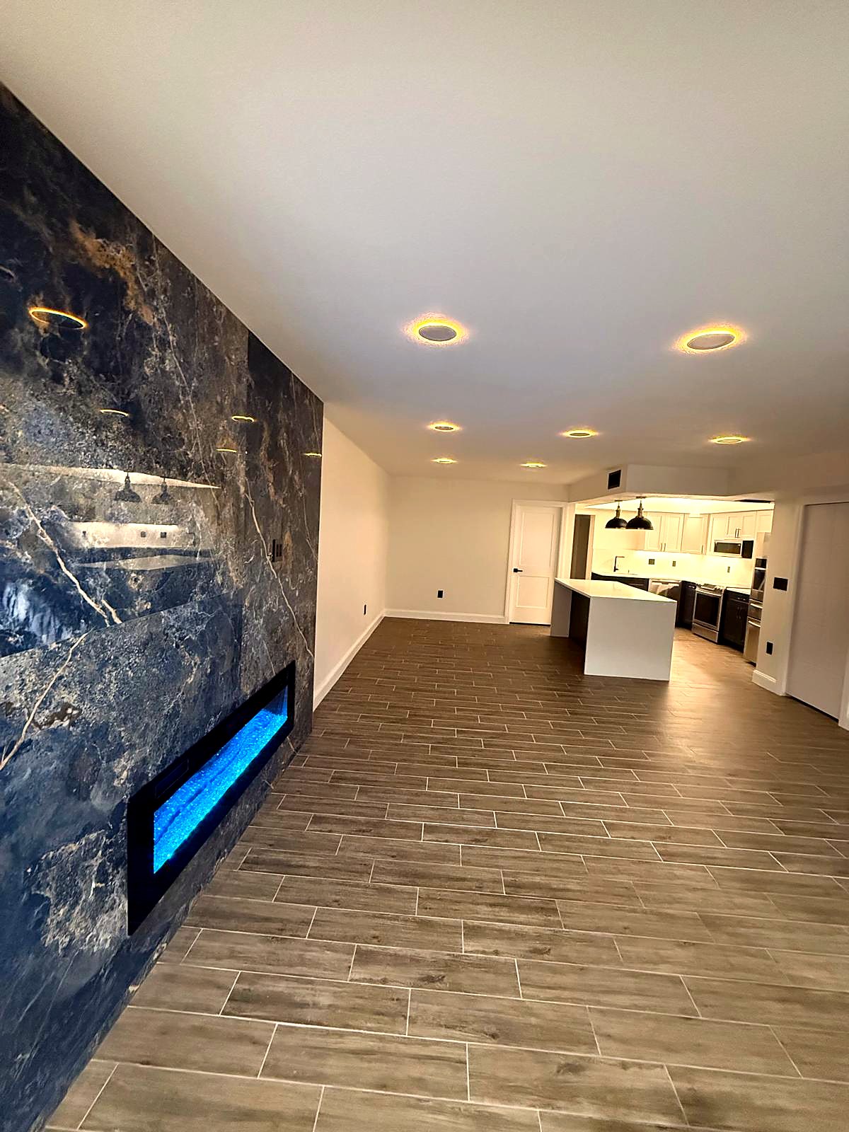 Looking along the slab wall and the lit fireplace toward the kitchen, with halo downlights across the ceiling