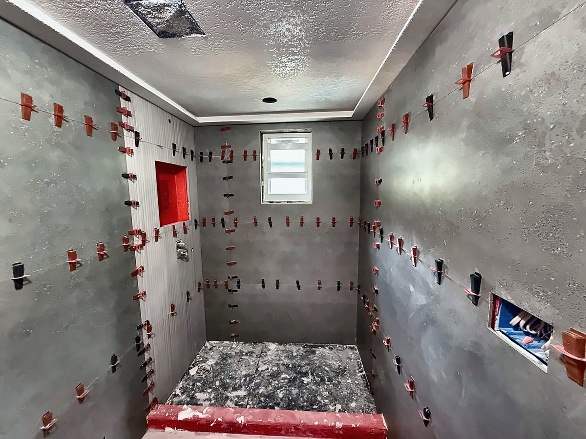 Shower walls being set in large-format tile with red leveling clips at every joint, the niche and curb already waterproofed