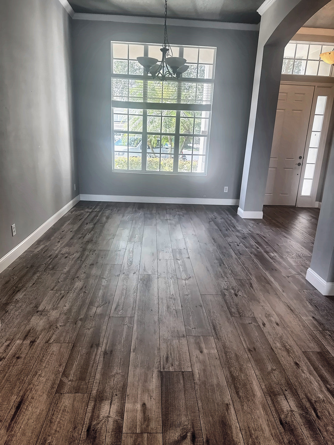 Dining room before renovation: an empty grey room with a tall multi-pane window, a wrought iron chandelier and an arched opening beside the front door