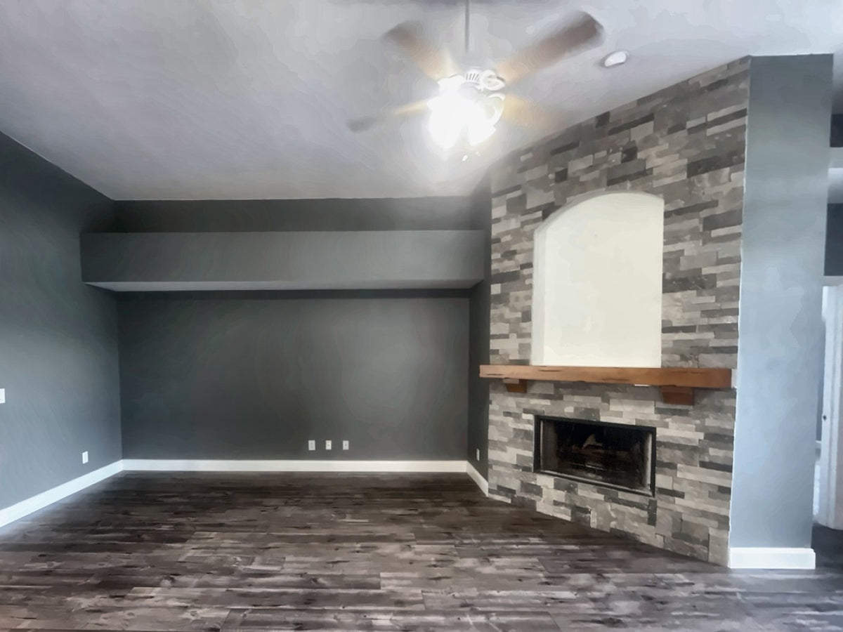 Great room before renovation: a stacked stone fireplace with an arched plaster niche and rustic timber mantel, under a dropped soffit, with gray walls and a ceiling fan