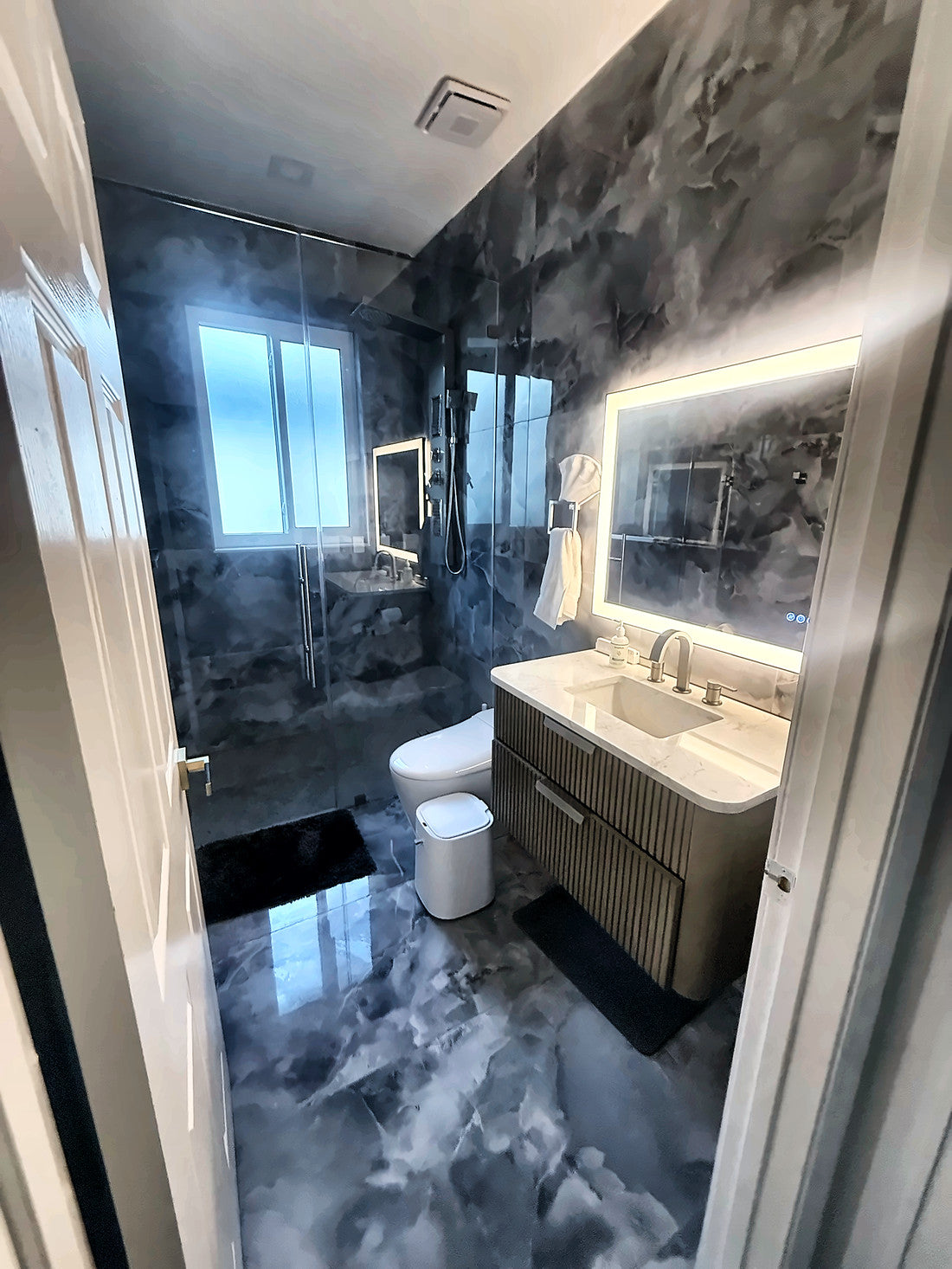 A second bath in dark onyx-look slab with a fluted floating vanity, backlit mirror and frameless shower