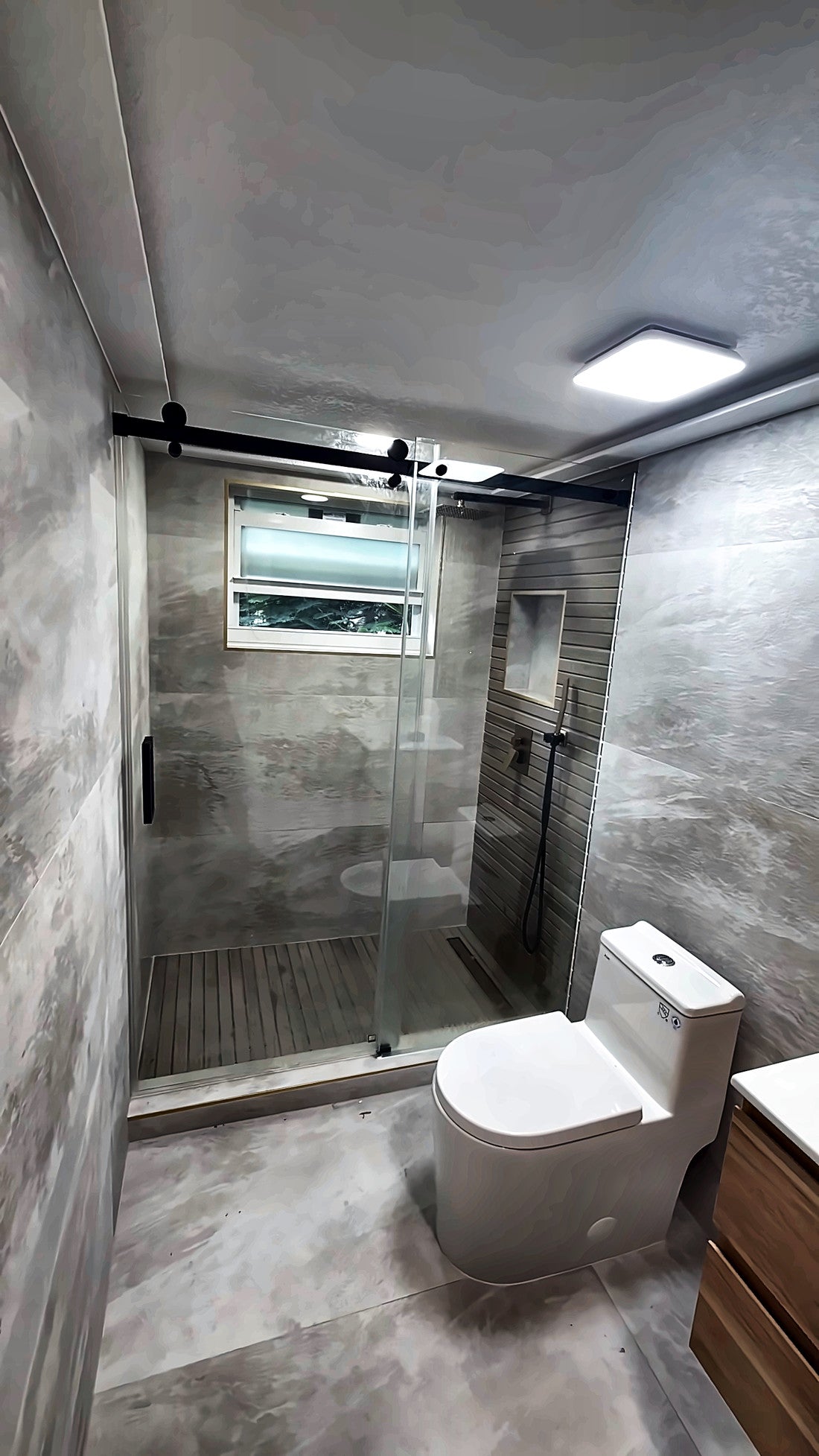 Walk-in shower behind a black-framed sliding glass door, grey stone-look tile on walls and floor and a window inside the shower