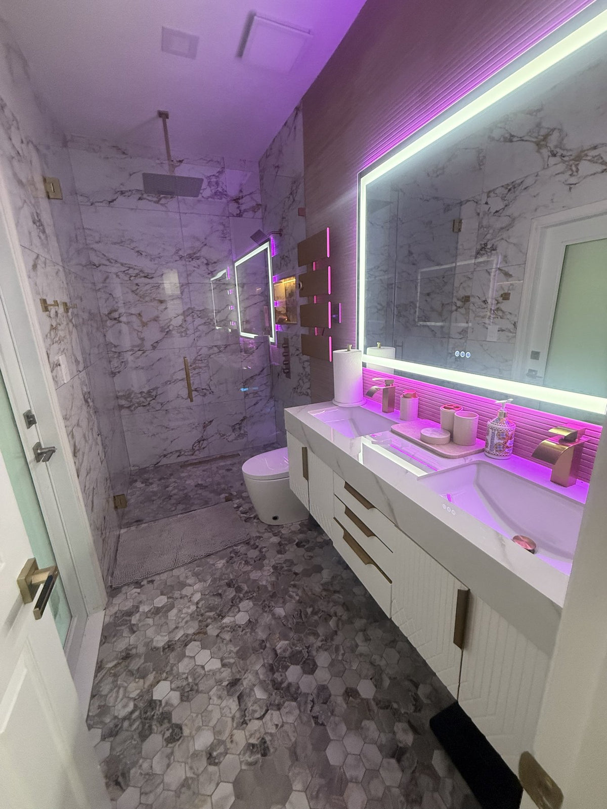 The same white and gold marble bath from the door: rain shower, lit niche, towel warmer and double vanity with waterfall faucets
