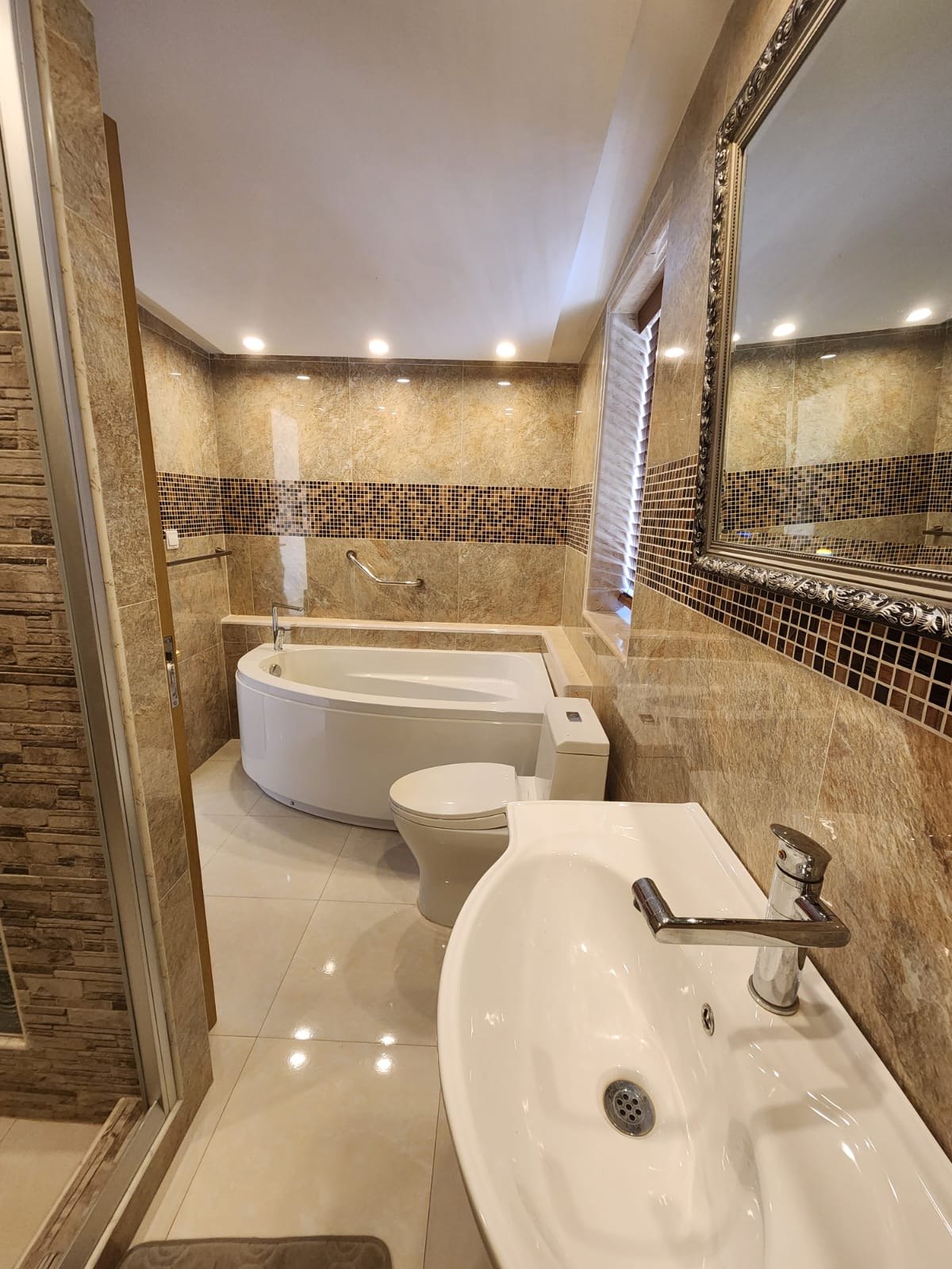 Bathroom in stone-look tile with a glass mosaic band, a corner tub, pedestal basin and recessed lights