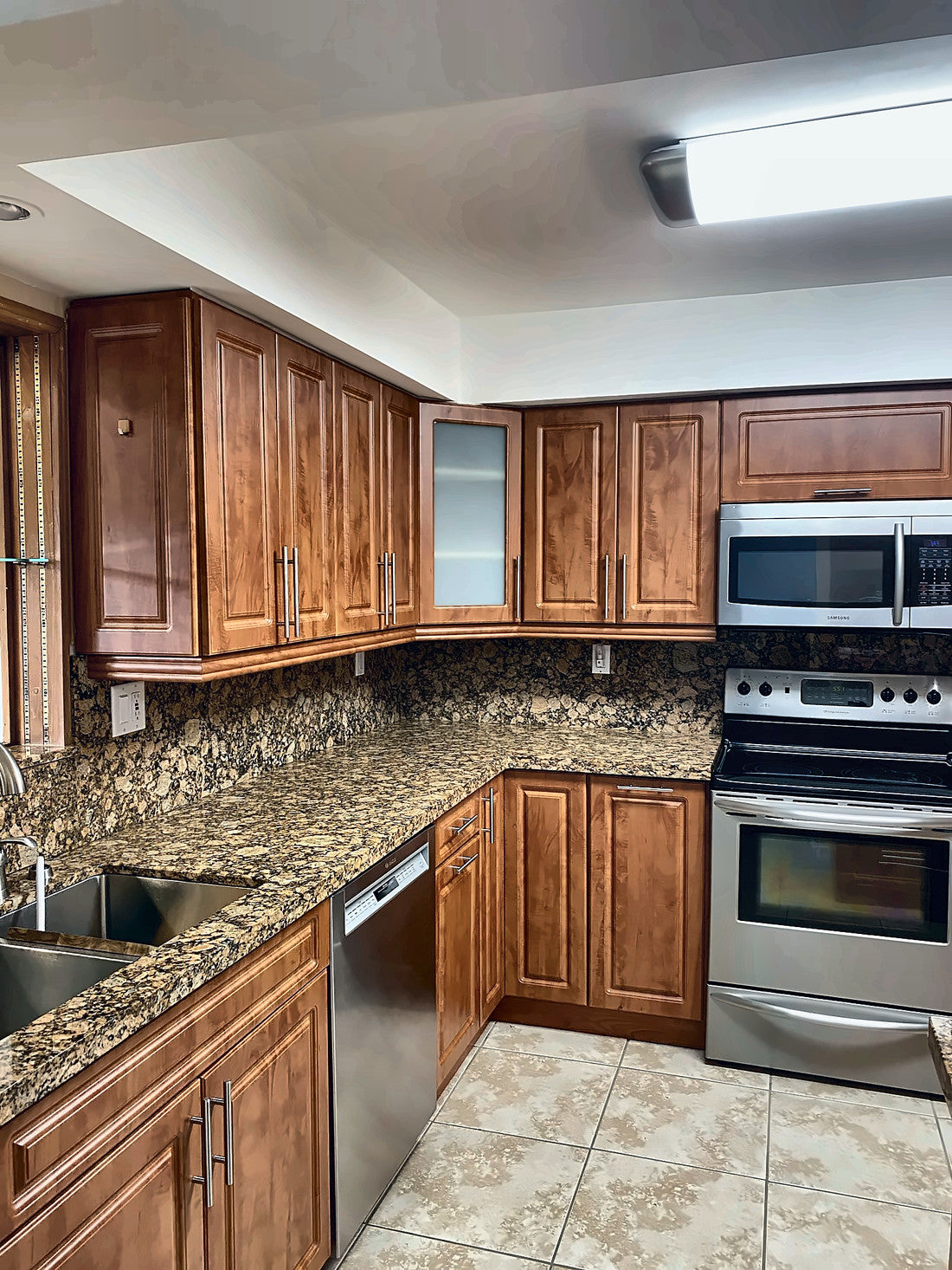 The kitchen before renovation: raised-panel cherry cabinets, speckled granite counters and backsplash, stainless appliances and beige ceramic floor under a boxed fluorescent light