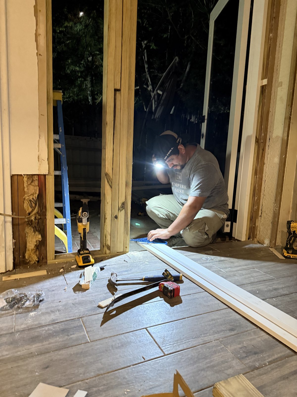 A worker in an Empire Nest shirt kneeling with a headlamp, measuring door trim after dark