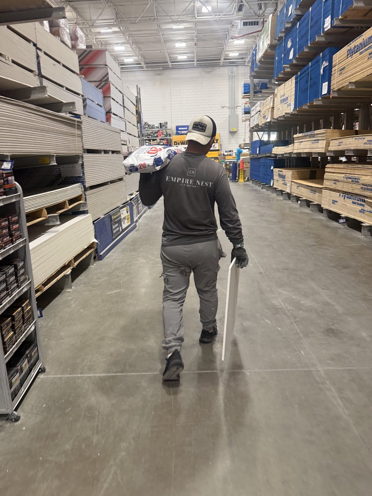 A worker in an Empire Nest shirt carrying a bag of concrete mix and a panel down a building supply aisle