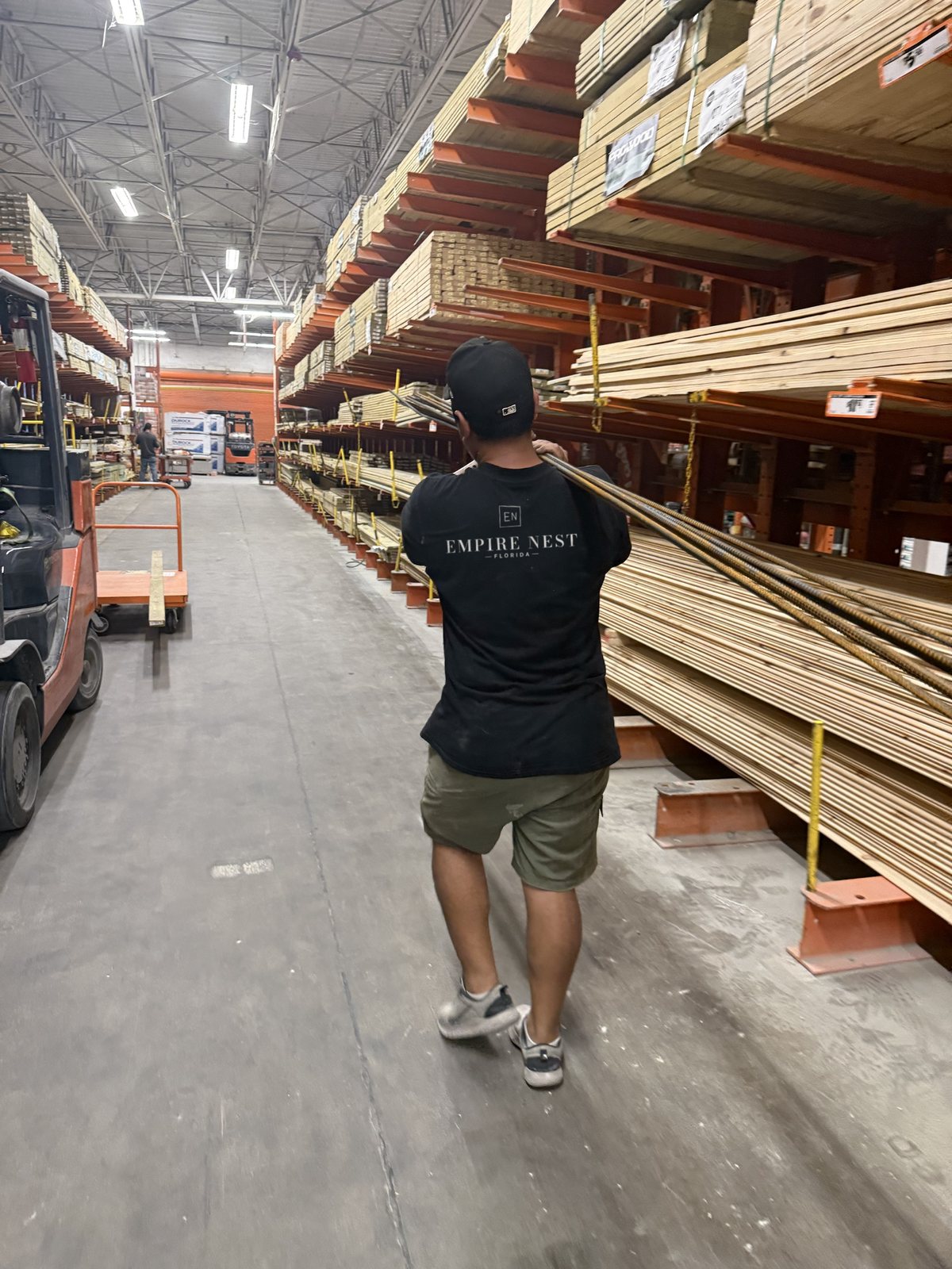A worker in an Empire Nest shirt carrying rebar on his shoulder down a lumber aisle