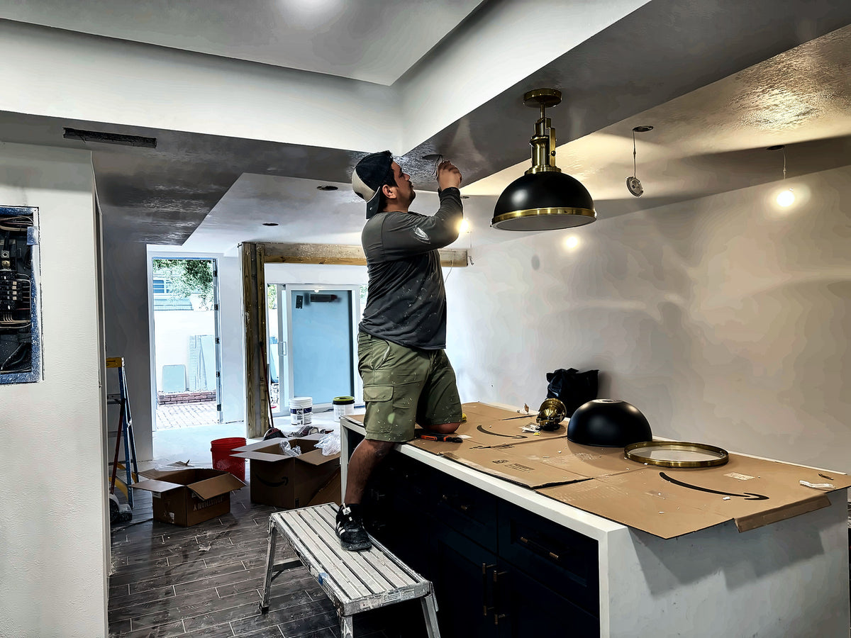 A tradesman wiring the brass and black pendants above the finished island, new sliding doors open behind